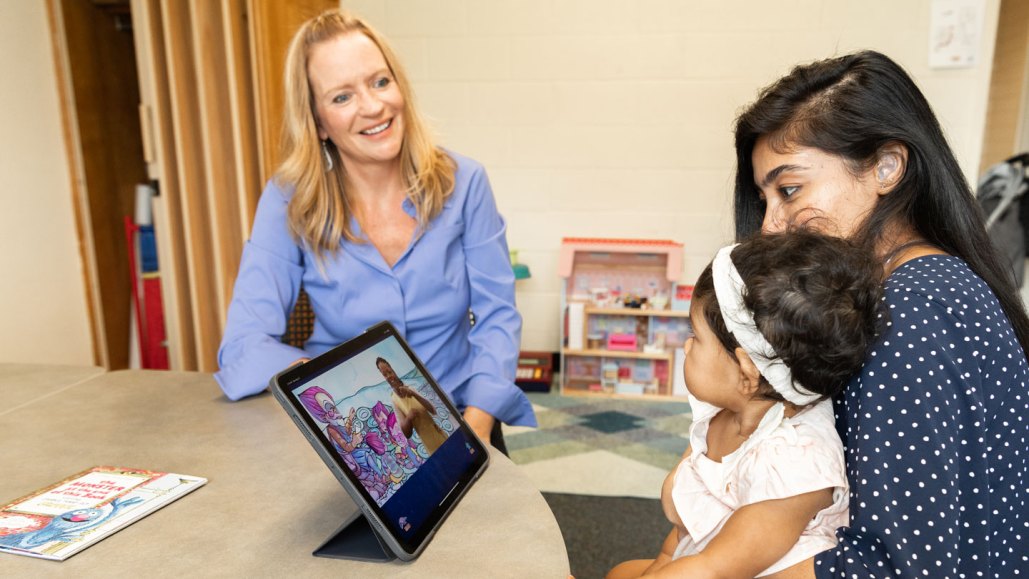 Rain Bosworth smiling and looking at a parent-child pair to her left. She has blonde hair and blue eyes and wearing blue button-up shirt. The parent is looking at an iPad, sitting in front of them on a round table. The iPad is displaying what appears to be a video with a person signing. The parent has black hair and wearing a navy polka dot shirt. The child is sitting on the parent's lap and staring at Bosworth.