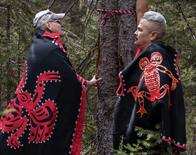 Two Indigenous men wear ceremonial black and red attire in front of a tree with red and black fabric tied around the trunk.